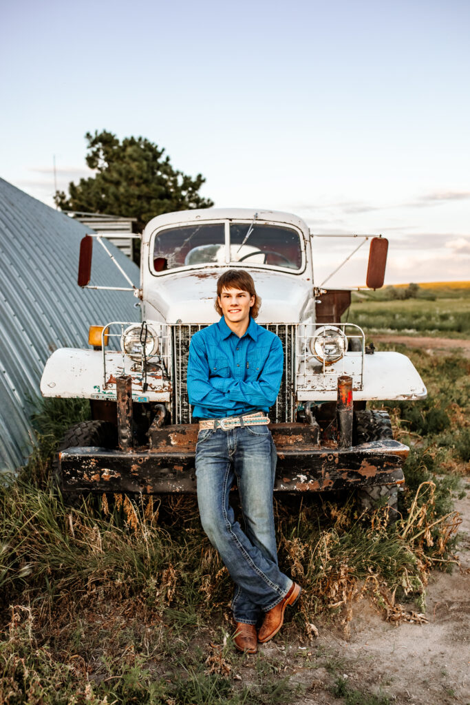 senior boy posing for senior portraits in jeans, cowboy pearl snap up and rodeo belt buckle