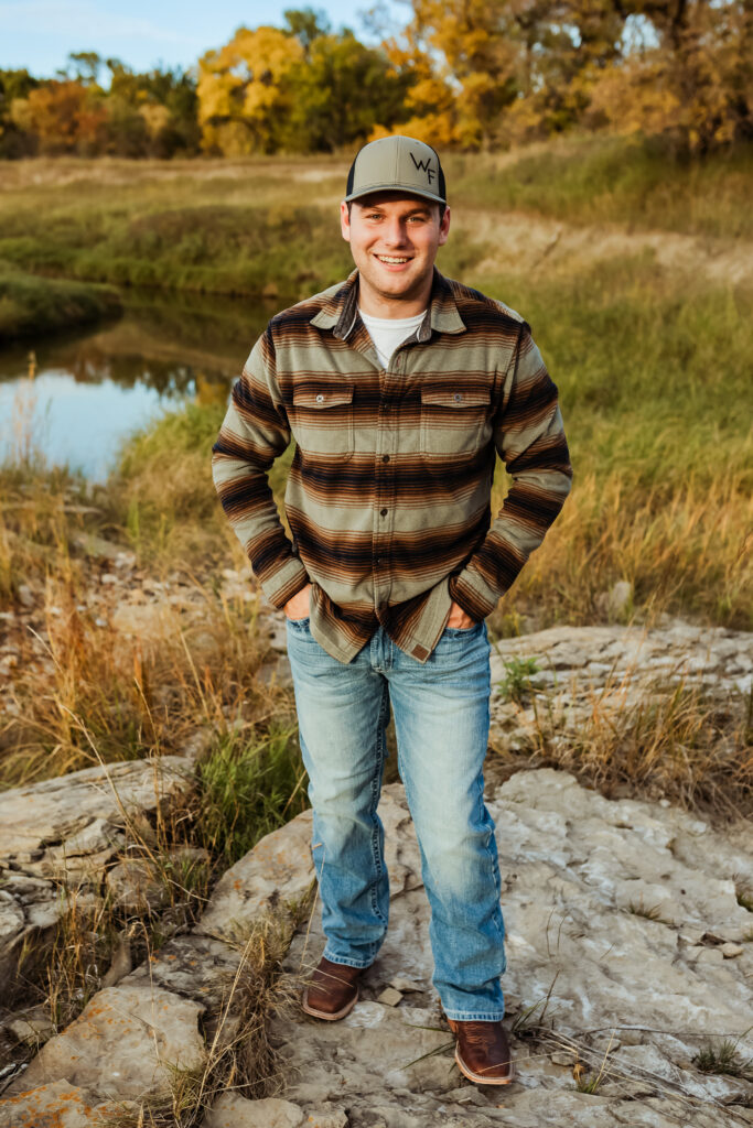 senior boy posing for senior portraits in field near a river in a flannel and jeans