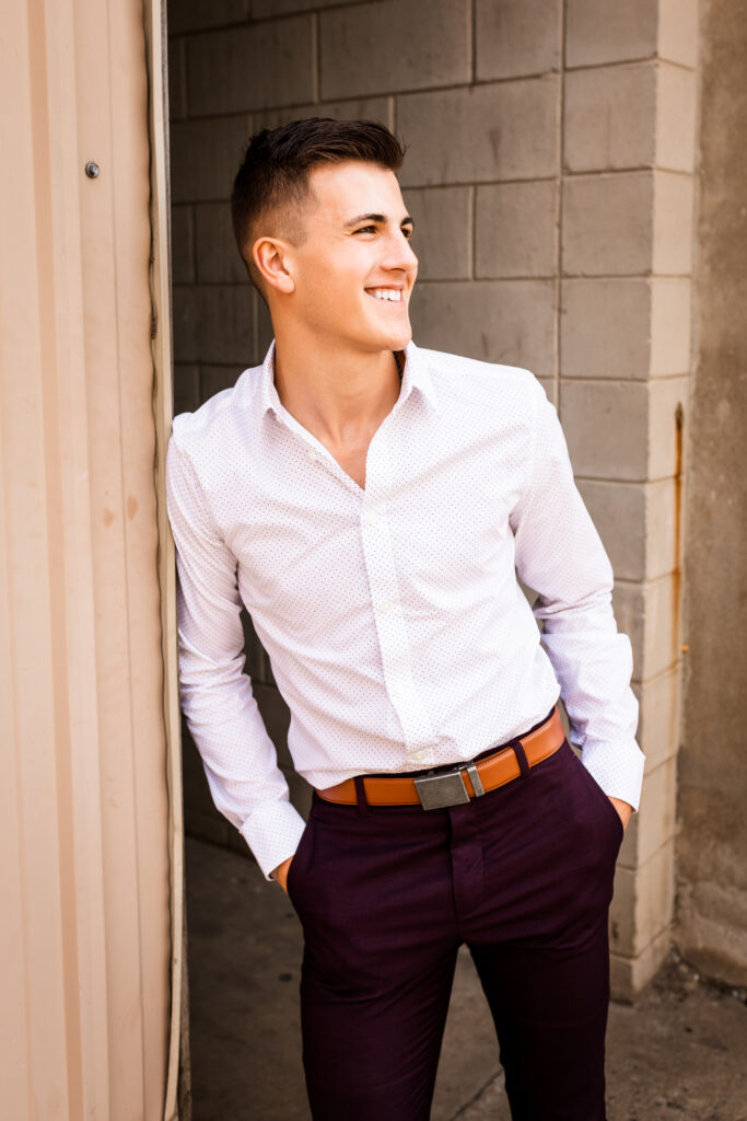 senior boy posing for senior portraits in an alley in a nice white button up and maroon pants with brown belt