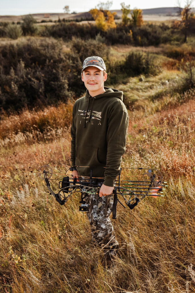 senior boy posing for senior portraits in sitka gear with bow in a field