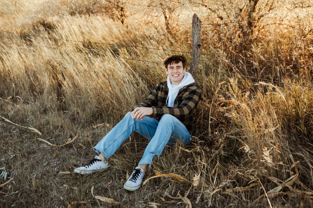 senior boy in a flannel shirt sitting near a fence post in a field