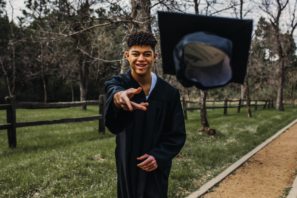 black senior boy posing for senior portraits in graduation gown tossing his cap towards the camera