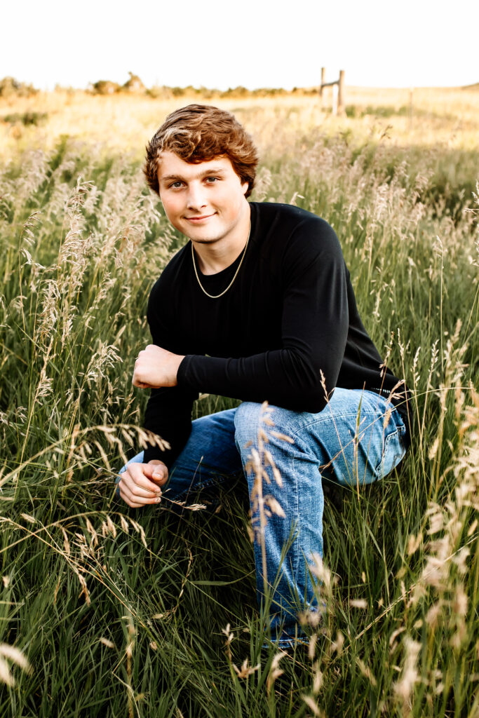 senior boy posing for senior portraits in a field with black long sleeve shirt and jeans